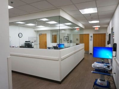 A clean, modern medical office with white walls, wood doors, computer stations, medical equipment, and bright ceiling lights.