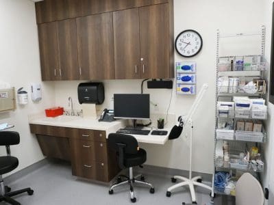 A medical exam room with a desk, computer, black chairs, wall clock, supply shelves, and various medical equipment and cabinets.