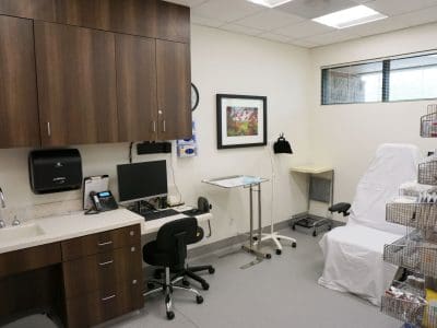A modern medical exam room with a desk, computer, sink, cabinets, exam chair covered in white sheet, and organized shelves with medical supplies.