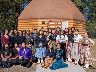 A group of young people in traditional and contemporary clothing pose in front of a log cabin structure on a sunny day.