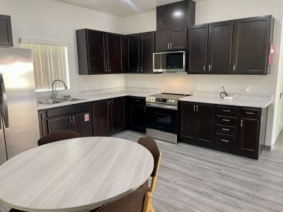 Modern kitchen with dark cabinets, stainless steel appliances, marble countertops, and a light wood dining table with four chairs on gray wood flooring.