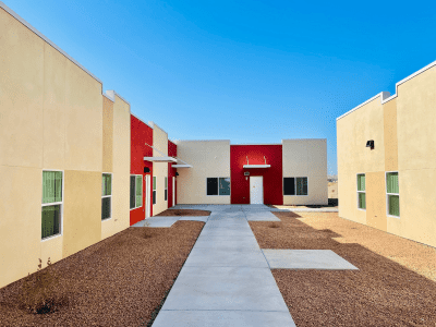 A modern, single-story building with red and beige walls and a concrete walkway, set against a clear blue sky and surrounded by gravel landscaping.