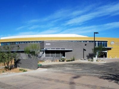 A modern, single-story building with gray and yellow walls, large windows, and desert landscaping in front, under a blue sky. The address 8880 is visible above the entrance.