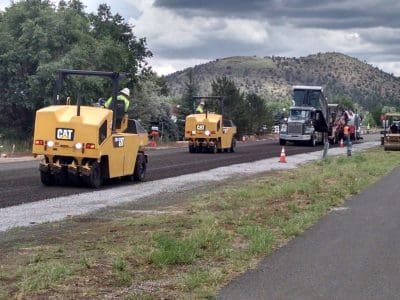 Construction workers operate yellow road rollers and an asphalt paver to pave a road, with trees and hills in the background under a cloudy sky.