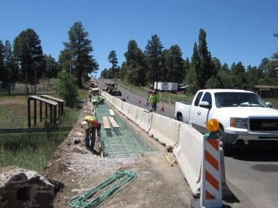 Workers construct a roadside pathway with concrete barriers separating the site from traffic; a white pickup truck and construction equipment are visible under clear skies.