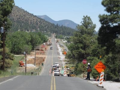 A rural road under construction with traffic cones, construction vehicles, and signs indicating "STOP" and "ROAD NARROWS." Trees and mountains are visible in the background.
