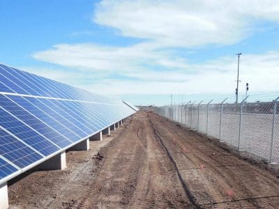 Rows of solar panels from the La Posada PV System are installed on the left side of a dirt pathway, with a chain-link fence running parallel on the right, under a partly cloudy sky.