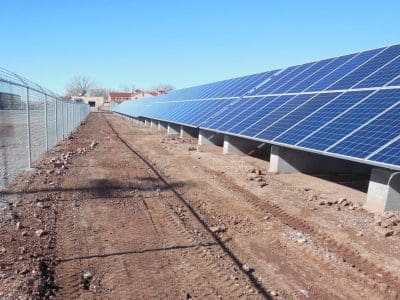 Rows of solar panels mounted on concrete bases beside a chain-link fence in a clear, open area with dirt and tire tracks visible on the ground; part of the La Posada PV System.