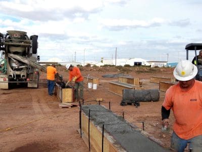 Construction workers pour and smooth concrete into wooden forms at the La Posada PV System construction site, with a cement mixer truck and a backhoe in the background.