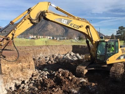 A yellow Caterpillar excavator is breaking rocks on a construction site, with mountains and trees in the background.