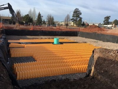 A construction site with yellow stormwater chambers installed in a gravel bed, surrounded by orange safety fencing and construction equipment.