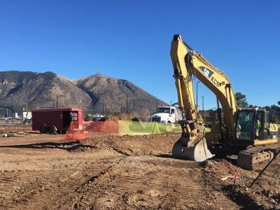 A construction site with a yellow excavator, a red trailer, piles of dirt, and a worker in a safety vest; mountains and a clear blue sky in the background.