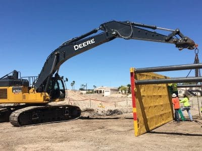 A large excavator lifts and positions a yellow trench shield while several construction workers in safety vests and helmets guide it at a worksite.
