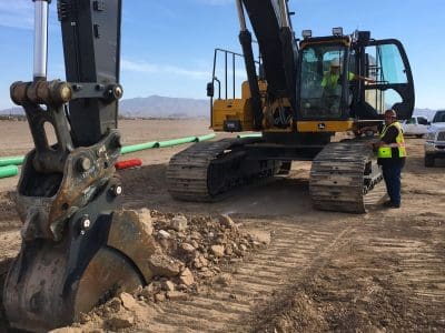A construction excavator on a dirt site with two workers in safety vests nearby and a white truck parked in the background.