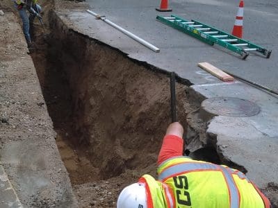 Two construction workers in safety gear dig a trench beside a street with nearby heavy machinery, traffic cones, and houses in the background.