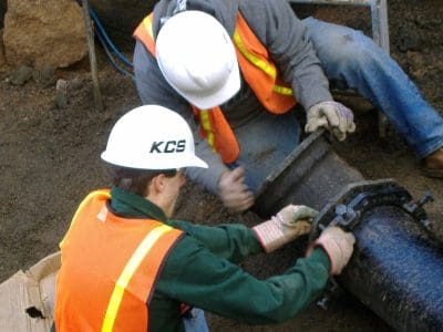 Two workers in safety vests and helmets repair a large underground pipe at a construction site.