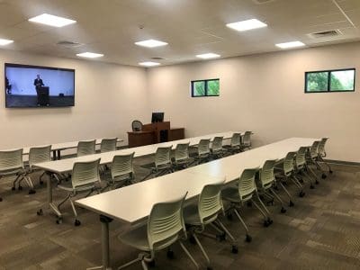 A classroom with rows of white tables and chairs, a podium and computer at the front, two small windows, and a large wall-mounted screen displaying a speaker.