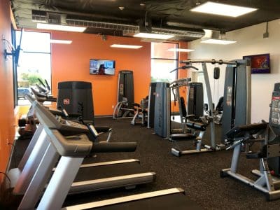 A gym room with treadmills, strength training machines, a stationary bike, orange accent walls, windows, and a TV mounted on the wall.