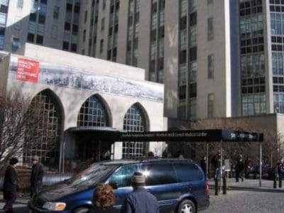 People walk outside the main entrance of NewYork-Presbyterian/Weill Cornell Medical Center, with a minivan parked in front of the building.