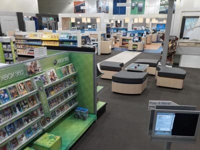 A retail electronics store interior with shelves displaying Xbox 360 games, demo stations, seating areas, and various technology products in the background.