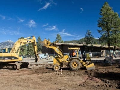 A construction site with a yellow excavator and a backhoe loader operating on dirt, surrounded by trees and distant buildings under a clear blue sky.