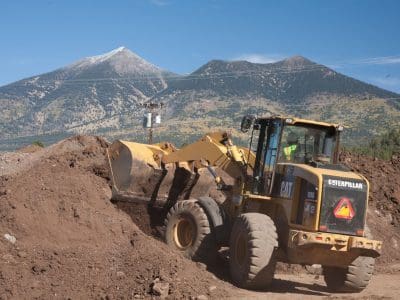 A yellow Caterpillar front loader moves dirt at a construction site with mountains and utility poles in the background.