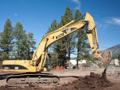 A yellow CAT excavator is digging and lifting soil at a construction site surrounded by trees and mountains under a clear blue sky.
