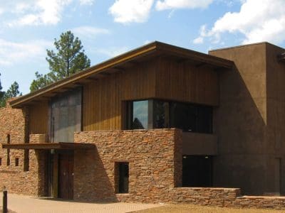 A modern building with stone and wood exterior walls, large windows, and a flat roof, surrounded by trees under a partly cloudy sky.