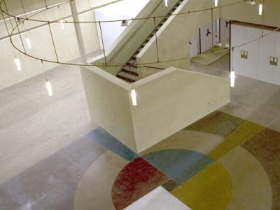 An industrial-style interior space with a geometric patterned floor, central staircase, exposed ceiling beams, and pendant lighting.