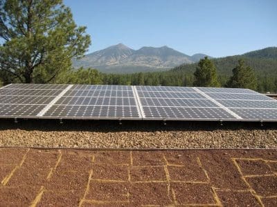 A row of solar panels, part of the Museum of Northern Arizona PV System, is installed on a flat, gravel-covered rooftop with trees and mountains visible in the background under a clear sky.