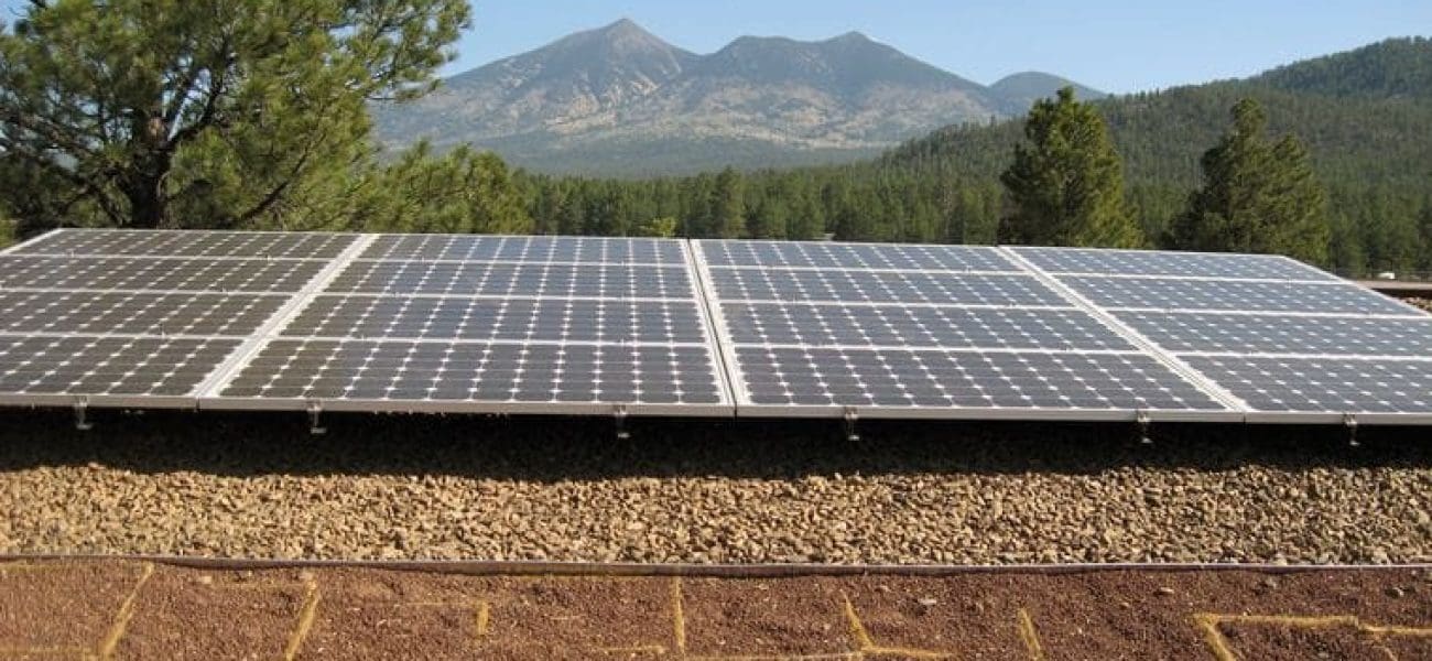 A row of solar panels, part of the Museum of Northern Arizona PV System, is installed on a flat, gravel-covered rooftop with trees and mountains visible in the background under a clear sky.