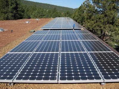 Rows of solar panels installed on a flat rooftop as part of the Museum of Northern Arizona PV System, with trees and hills visible in the background.