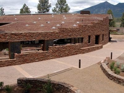 A modern building with stone walls and large windows sits amid landscaped pathways and trees, with mountains in the background—home to the Museum of Northern Arizona PV System.
