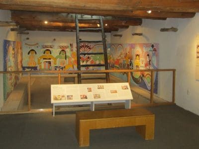 A museum exhibit room with a bench, informational panels, and a brightly colored mural on the far wall, bordered by wooden beams and a ceiling with exposed logs.