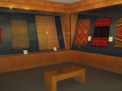 A museum exhibit displays several patterned woven textiles hanging on blue walls behind glass, with descriptive placards and a wooden bench in the center of the room.