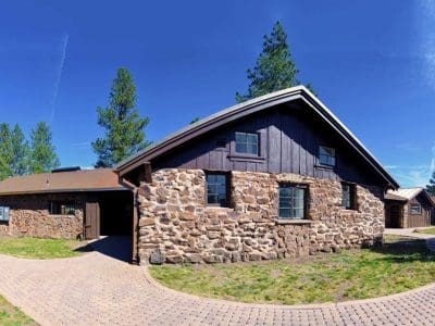 Single-story stone and wood house with a sloped roof, surrounded by a paved driveway and green lawn, set against a clear blue sky and tall trees.