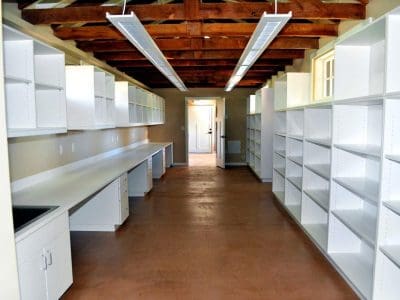 Room with exposed beam ceiling, fluorescent lighting, white countertops, and empty white shelving units lining both walls, leading to a door at the far end.
