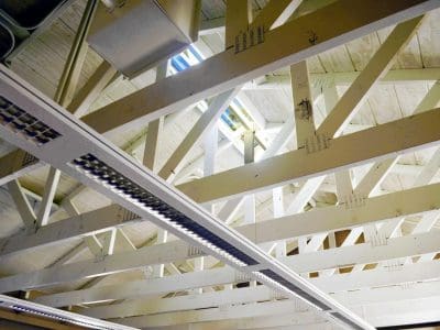 Exposed white wooden roof trusses and beams with fluorescent light fixtures and a small skylight above.