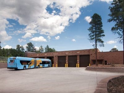 A blue city bus labeled "Grand Canyon National Park" approaches a brown garage building surrounded by trees on a clear day.