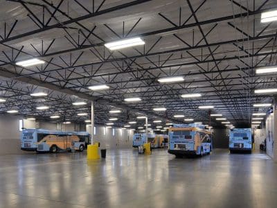 A large, well-lit indoor garage houses five buses parked in rows on a polished concrete floor under a high, open ceiling.