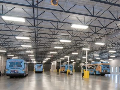 Several blue and yellow public buses are parked inside a large, well-lit indoor garage with high ceilings and exposed beams.