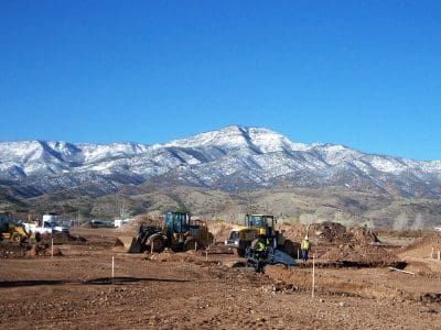Construction vehicles and workers operate on a dirt site with a snow-capped mountain range in the background under a clear blue sky.
