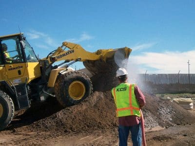 A construction worker operates a Komatsu front loader, dumping soil onto a pile, while another worker in a high-visibility vest and hard hat observes.