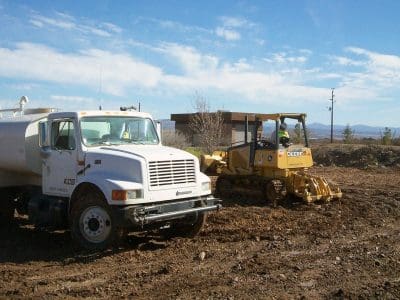 A water truck sprays the ground while a bulldozer levels dirt at a construction site under a clear sky.