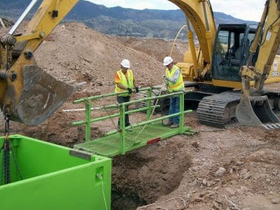 Two construction workers in safety gear stand on a metal platform over a trench, working with an excavator at a dirt construction site.