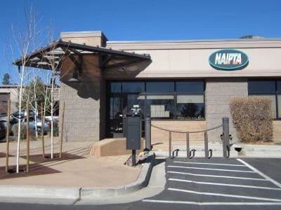 Single-story NAIPTA office building with large windows, a small awning over the entrance, a few leafless trees, and an accessible parking space in front.