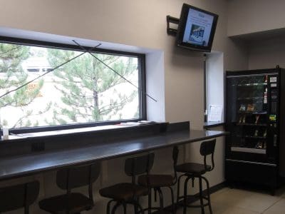 Break room with a long counter, stools, a vending machine, two microwaves, a window with trees outside, and a wall-mounted TV displaying information.