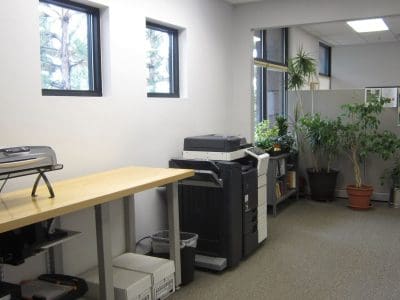 Office break room with a countertop toaster oven, a copier machine, potted plants, and bulletin boards on the wall by windows and a partition.