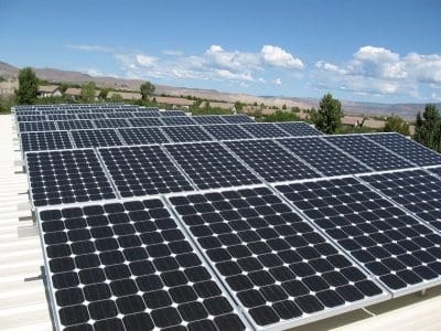 Rows of solar panels installed on a flat rooftop at the Mountain Line Verde Valley Transit Facility PV System under a clear blue sky, with scattered clouds and distant hills in the background.