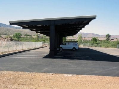 A white van is parked under the Mountain Line Verde Valley Transit Facility PV System’s large metal carport structure in an otherwise empty asphalt parking lot, with mountains and houses visible in the background.